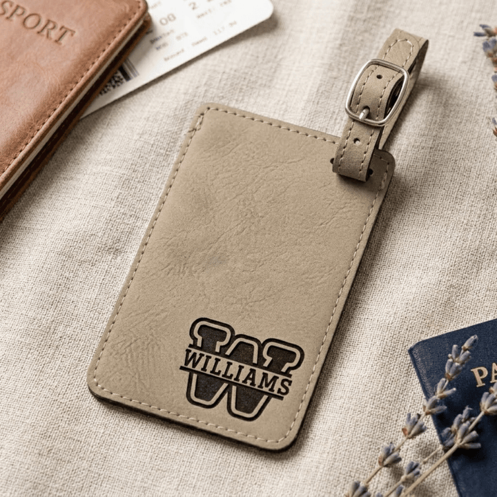 Beige leather luggage tag with 'Williams' logo engraved in bottom right corner laying on a textured surface with travel documents.