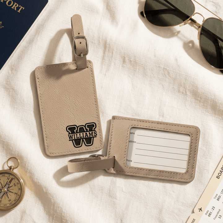 Beige leather luggage tags with front showing 'WILLIAMS' monogram engraved in black and the back of the tag with a clear plastic opening to put your personal information.