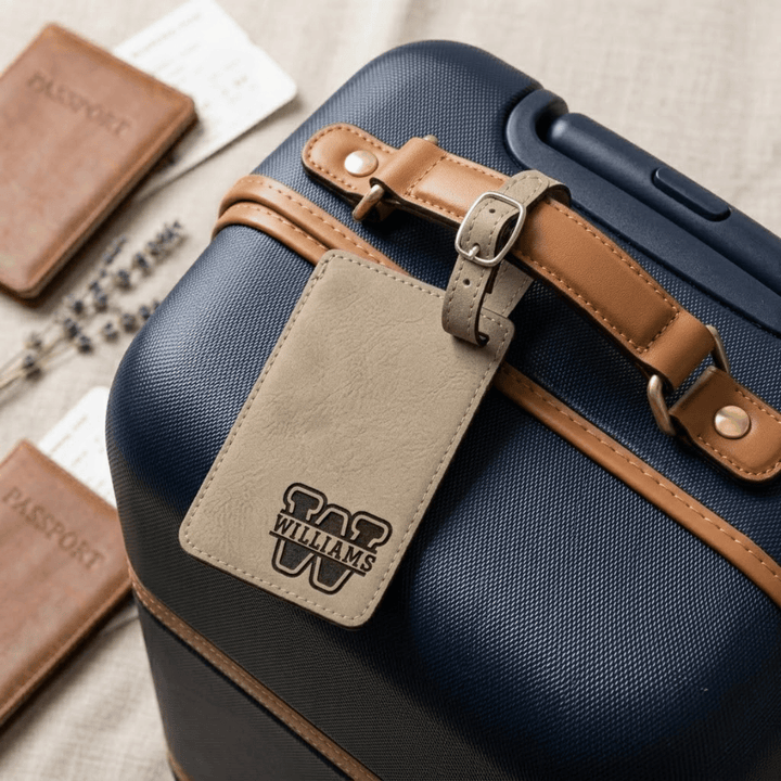 Blue suitcase with a brown strap and beige tag featuring 'Williams' varsity split monogram, surrounded by passports on a textured surface.