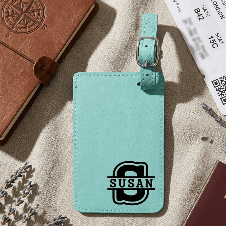 Teal luggage tag with 'SUSAN' varsity style split monogram engraved in black in bottom right corner of tag laying on a textured surface with travel items.