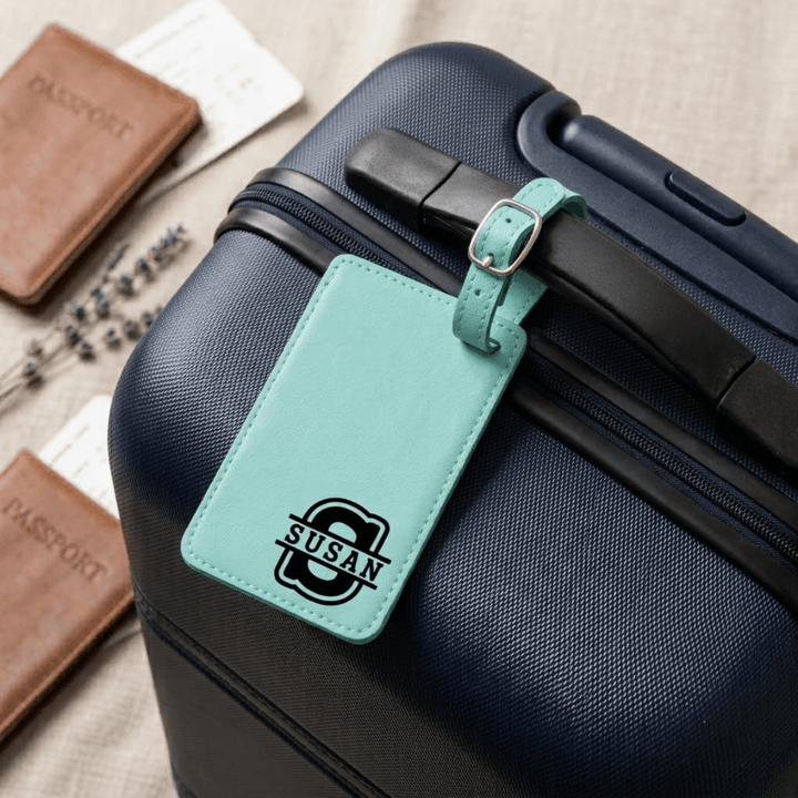 Teal luggage tag with 'SUSAN'  monogram buckled on a navy suitcase with passports and lavender flowers in the background.