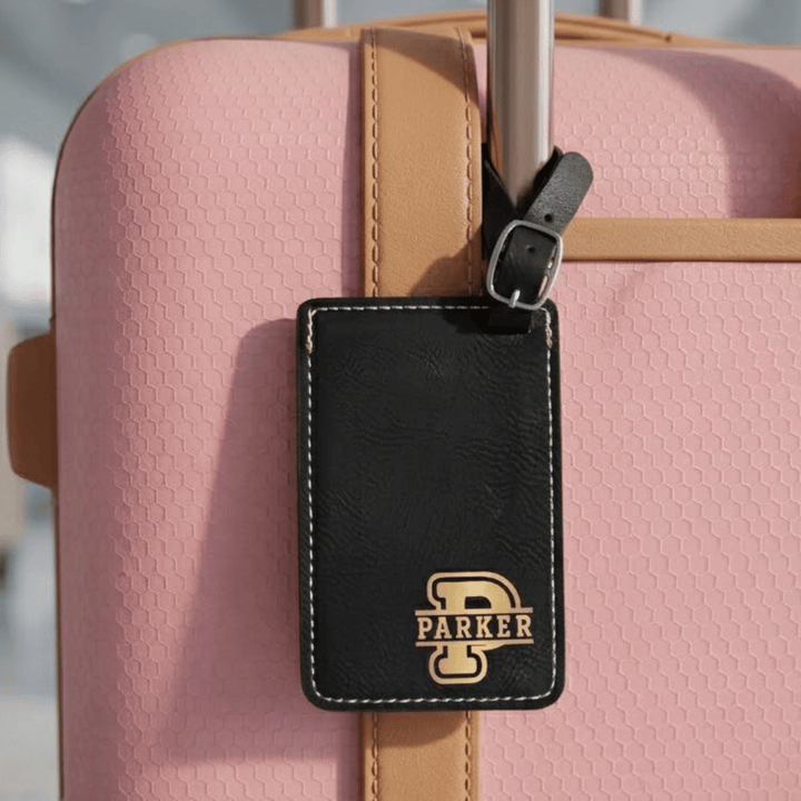 Black luggage tag with 'PARKER' monogram engraved in metallic gold in bottom right corner. Tag is buckled on a pink suitcase
