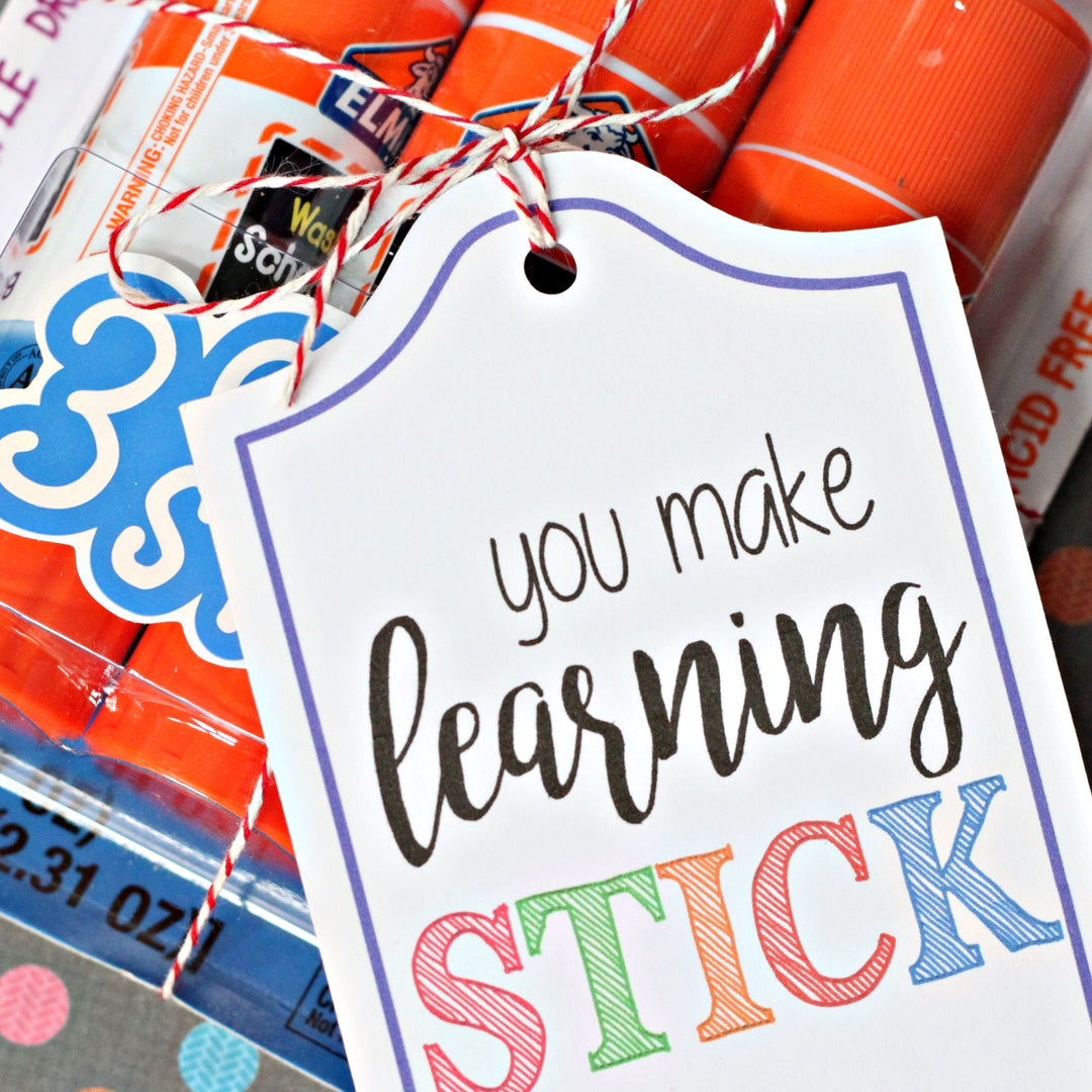 A close-up of a gift tag tied with red-and-white string, reading 'you make learning STICK' in black and multicolor lettering, with orange and white glue sticks and craft supplies visible in the background.