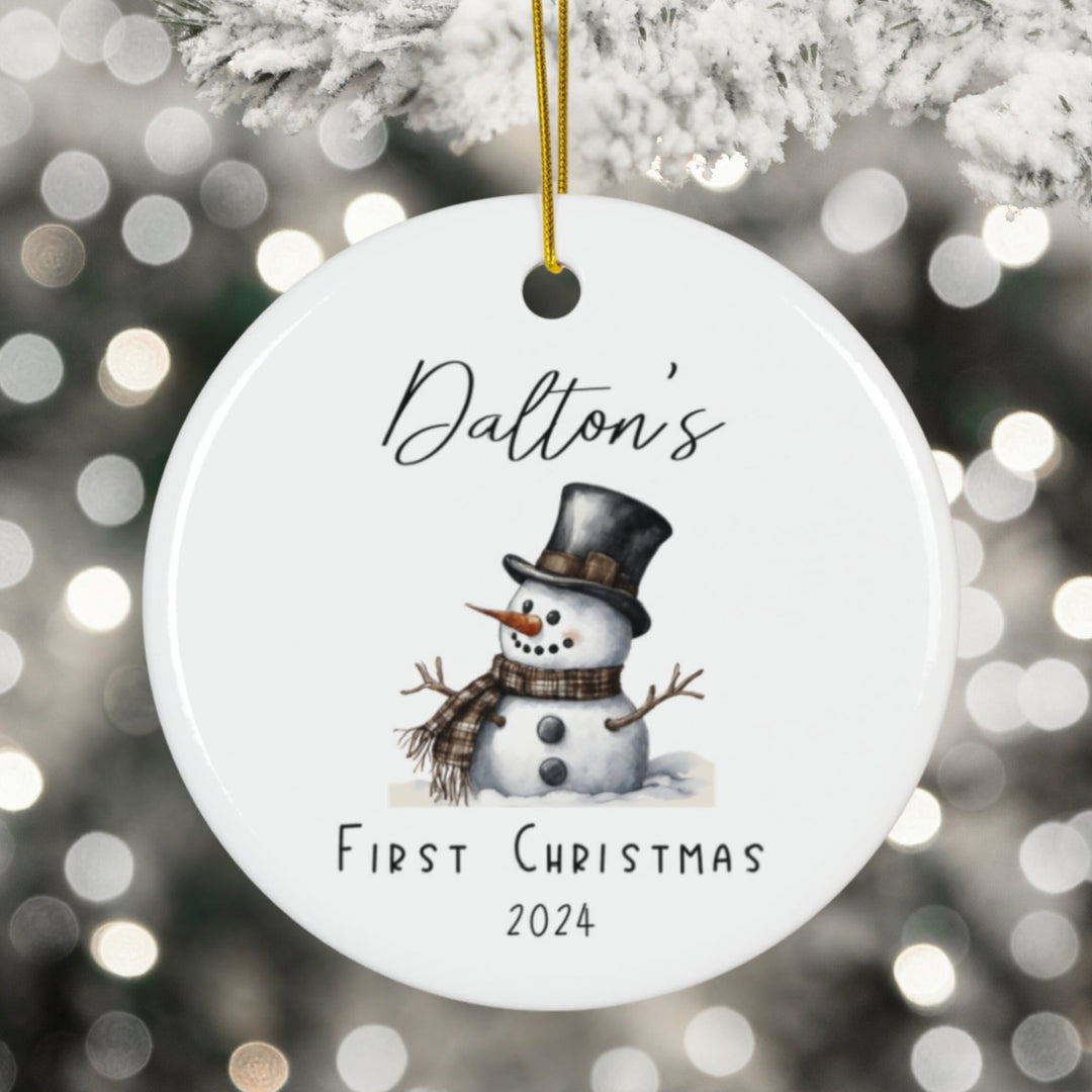 Round Christmas ornament with a snowman design and text 'Dalton's First Christmas 2024' against a blurred festive background.