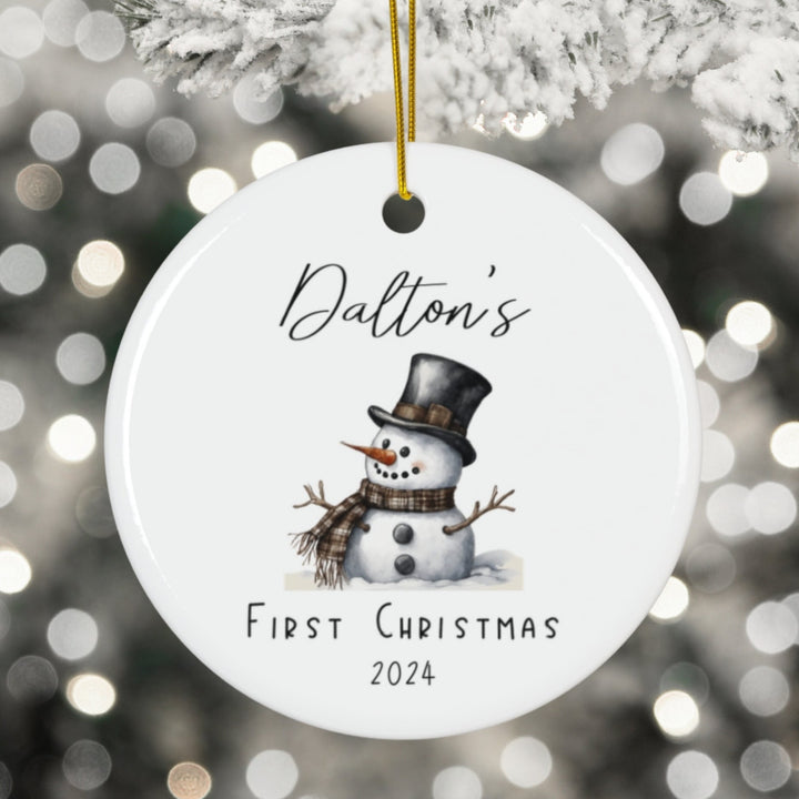 Round Christmas ornament with a snowman design and text 'Dalton's First Christmas 2024' against a blurred festive background.
