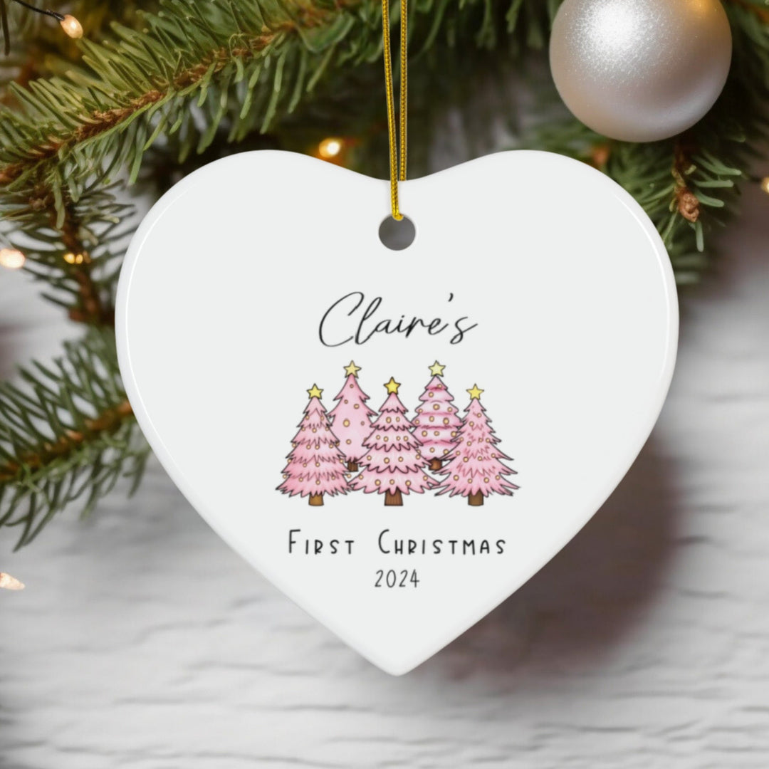 Heart-shaped ceramic Christmas ornament with pink Christmas trees and text that says 'Claire's First Christmas' and the year,  hanging on a tree.