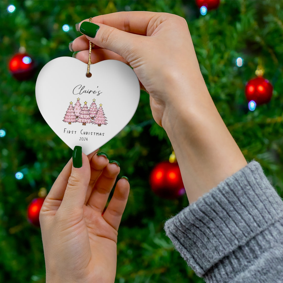 Heart-shaped ornament with pink Christmas trees and 'Claire's First Christmas' text along with the year, held against a Christmas tree background.