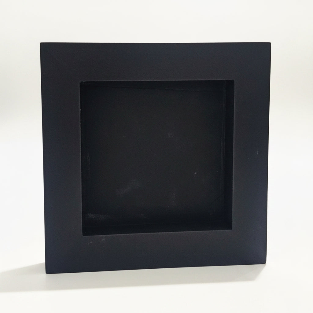 A square black back of a 6x6 inch wood sign with a recessed center, standing on a white surface with a blurred indoor background.