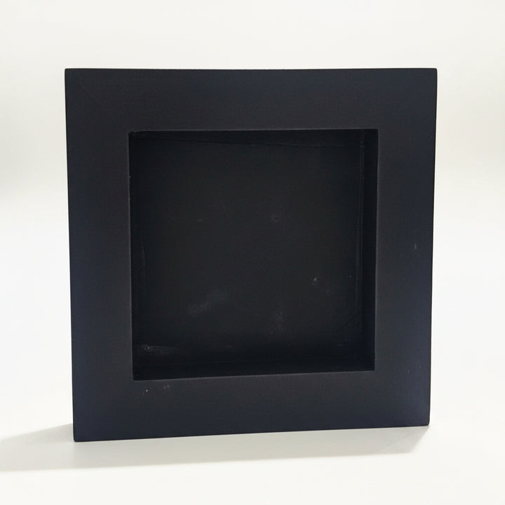 A square black back of a 6x6 inch wood sign with a recessed center, standing on a white surface with a blurred indoor background.