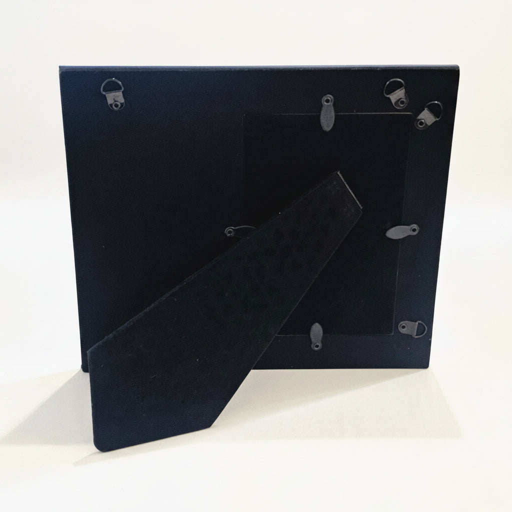 The back view of a picture frame with metal mounting clips and a hinged support brace, standing on a white surface.