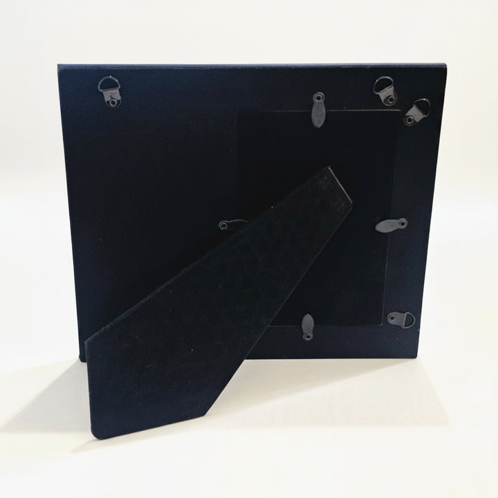 The back view of a picture frame with metal mounting clips and a hinged support brace, standing on a white surface.