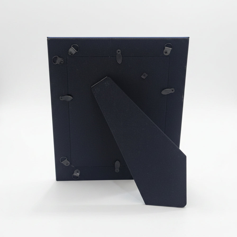 The back of a black picture frame with metal hanging clips and a fold-out leg, standing on a white tabletop in an indoor room setting.