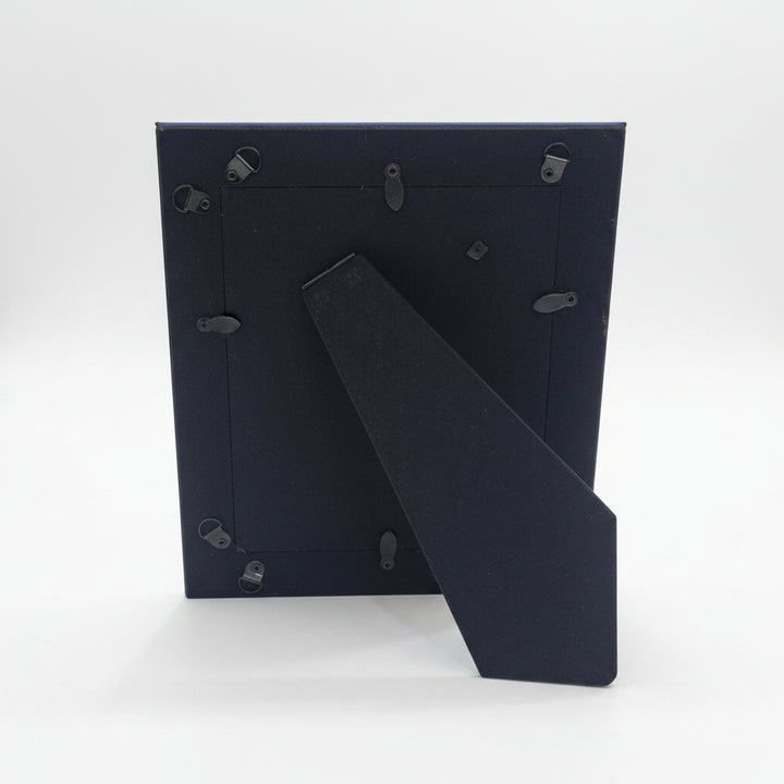 The back of a black picture frame with metal hanging clips and a fold-out leg, standing on a white tabletop in an indoor room setting.