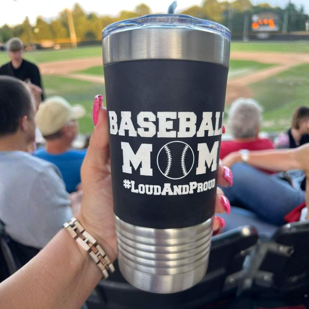 Person holding a 'Baseball Mom LoudAndProud' tumbler at a baseball game.