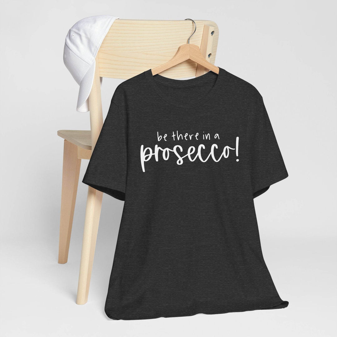 Dark heather gray t-shirt hanging on a hanger with 'be there in a prosecco!' text on a wooden chair against a white background