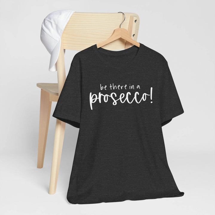 Dark heather gray t-shirt hanging on a hanger with 'be there in a prosecco!' text on a wooden chair against a white background