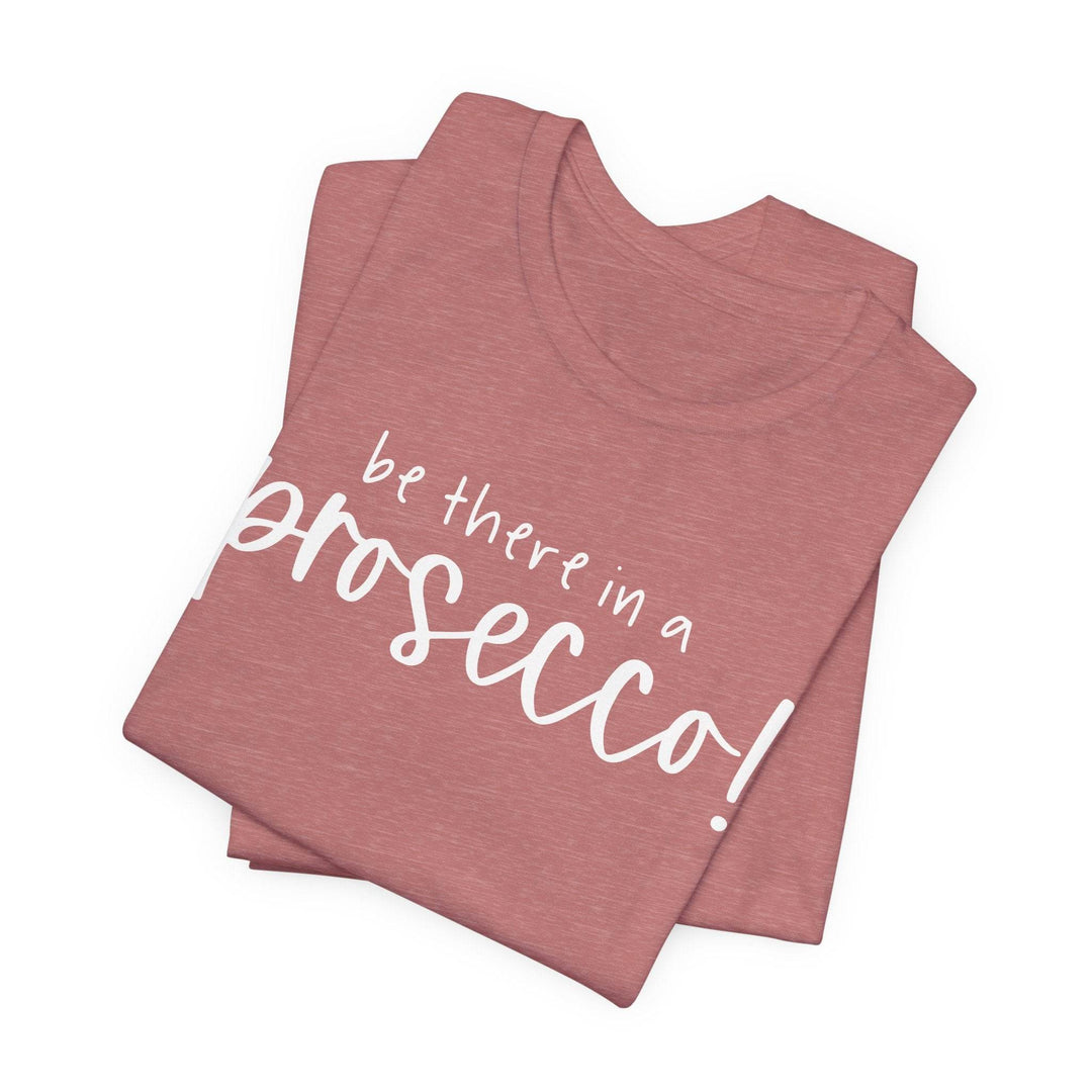 Stack of folded heather mauve t-shirts with 'be there in a prosecco!' text on a white background
