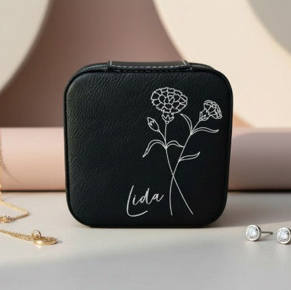 Black travel jewelry box with birth month flower design and 'Lida' name engraved in metallic silver in a modern script font surrounded by a necklace and earrings on a neutral background.