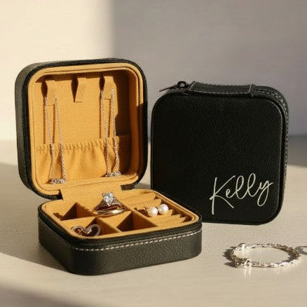 Black jewelry case with 'Kelly' engraved in metallic silver, opened to reveal jewelry on a beige surface