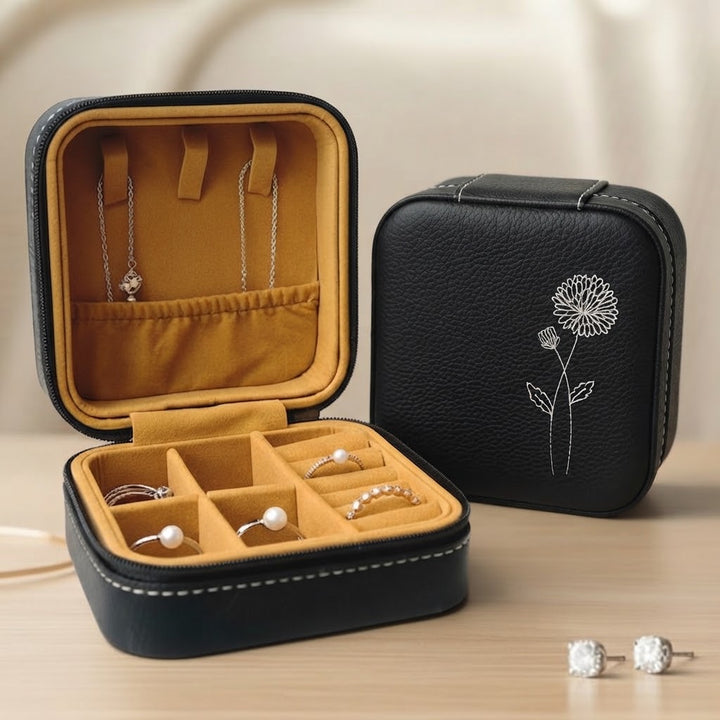 A black faux-leather jewelry organizer case shown open and closed on a light wood surface. The open case has a mustard-yellow interior with ring slots, a necklace loop, and small pearl and silver rings displayed inside.