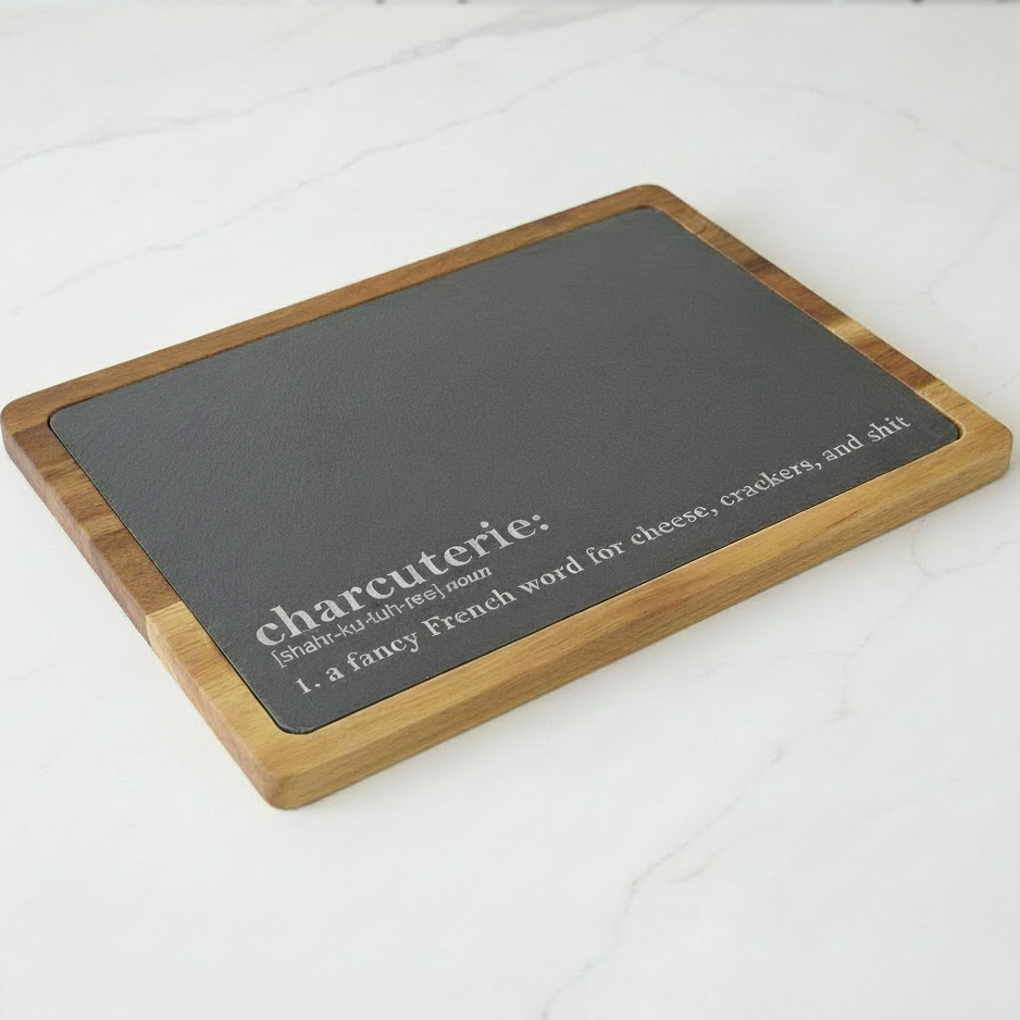 Rectangular charcuterie board with black slate surface and acacia wood frame on a white surface. Funny definition of charcuterie board is engraved on slate. It says, 'a fancy French word for cheese, crackers, and shit'