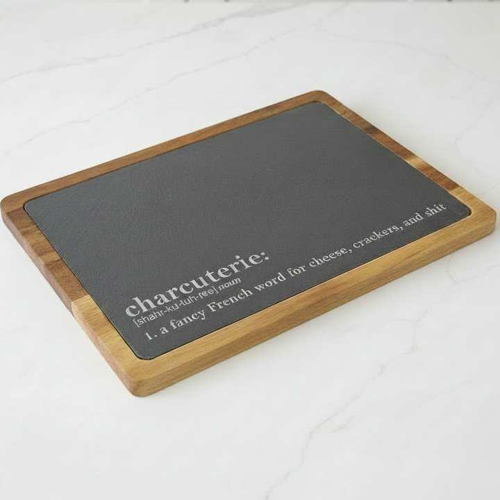 Rectangular charcuterie board with black slate surface and acacia wood frame on a white surface. Funny definition of charcuterie board is engraved on slate. It says, 'a fancy French word for cheese, crackers, and shit'