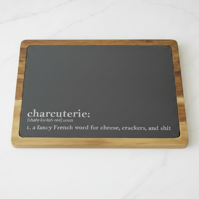 Rectangular cutting board with slate surface and acacia wood frame on a white kitchen counter. On the bottom of the slate is the funny definition of charcuterie board in the style of a dictionary definition. The definition says, 'a fancy French word for cheese, crackers, and shit'
