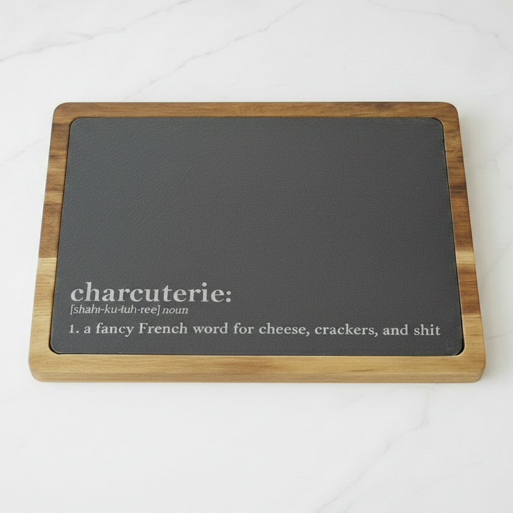 Rectangular cutting board with slate surface and acacia wood frame on a white kitchen counter. On the bottom of the slate is the funny definition of charcuterie board in the style of a dictionary definition. The definition says, 'a fancy French word for cheese, crackers, and shit'