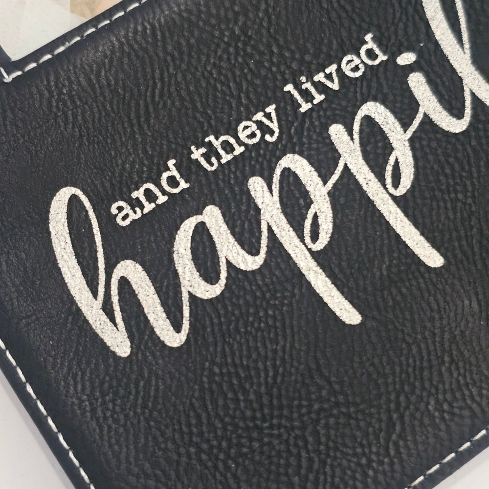 Close-up of a black faux-leather picture frame with white stitching and the phrase 'and they lived happily ever after' in metallic silver lettering.