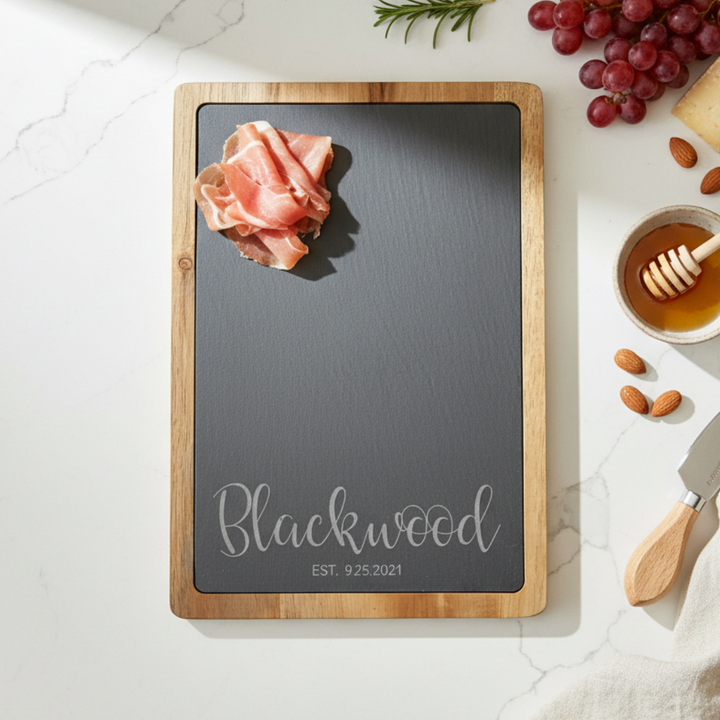 Wood and slate charcuterie board with 'Blackwood' engraved in slate on a marble surface with food items.