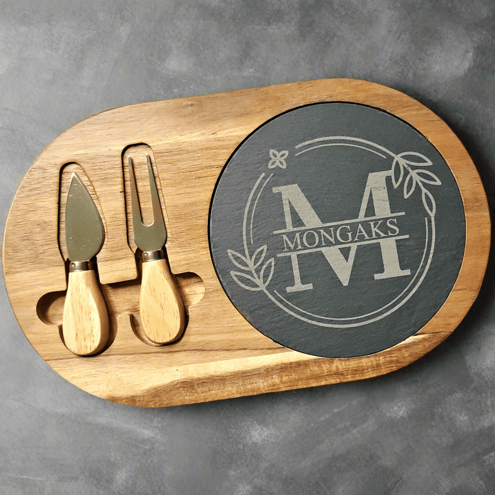 Wooden cutting board with cheese knives and a round slate piece engraved with and initial and family name sitting on a gray background