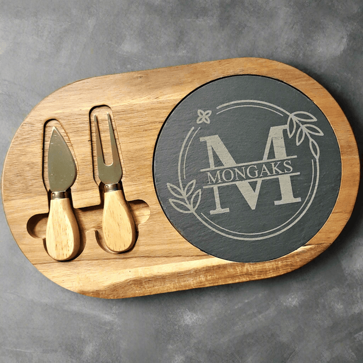 Wooden cutting board with cheese knives and a round slate piece engraved with and initial and family name sitting on a gray background