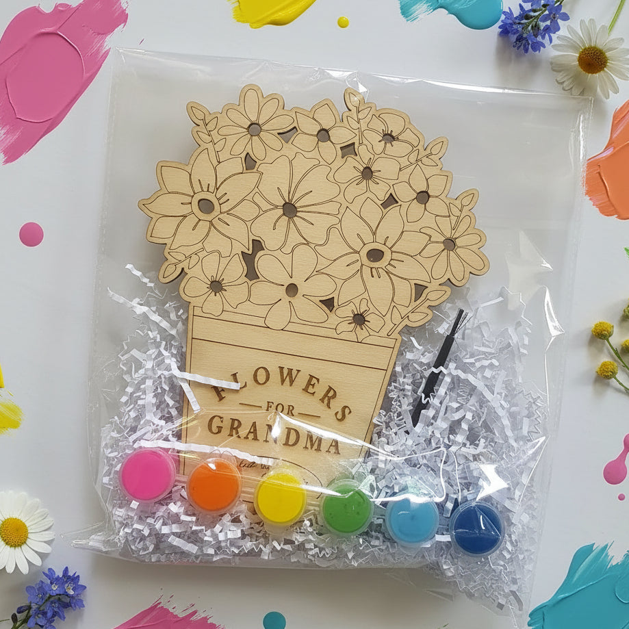 A floral gift set in a clear plastic bag with shredded white paper. The set includes a wooden flower pot design engraved with 'FLOWERS FOR GRANDMA,' several small round buttons in pink, orange, yellow, green, light blue, and dark blue, and a black hair pin.