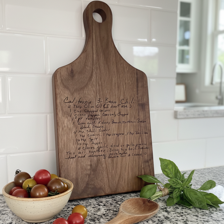 Wooden cutting board with engraved handwritten recipe on a kitchen counter with tomatoes and basil.