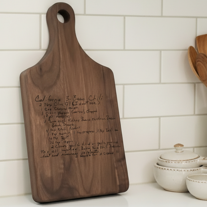 Wooden cutting board with engraved handwritten recipe on a kitchen counter with white tiled wall and utensils.
