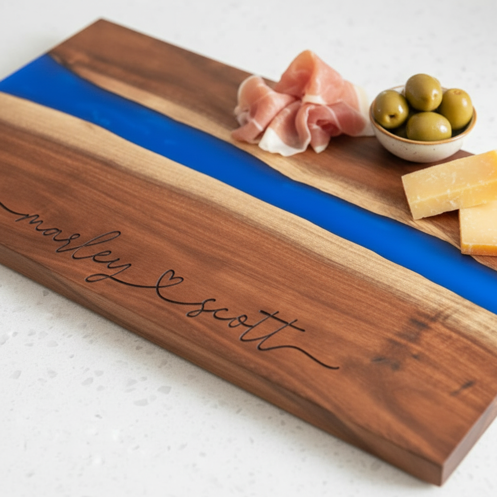Wooden charcuterie board with blue resin inlay, prosciutto, olives, and cheese. Two names connected with a heart in the middle are engraved on bottom of board.