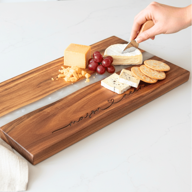 Wooden cheese board with engraved names in a modern script font connected with a heart and assorted cheeses, fruits, and crackers on a kitchen counter.