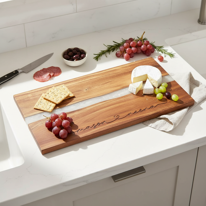 Wooden charcuterie board with 2 engraved names connected with a heart in a modern script, with cheese and grapes on a kitchen counter