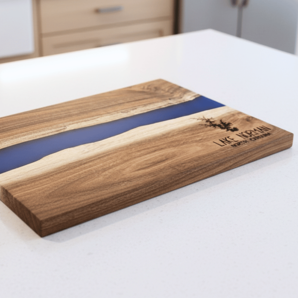 Wooden cutting board with blue resin inlay on a white surface and custom lake name and lake silhouette engraved in bottom right corner