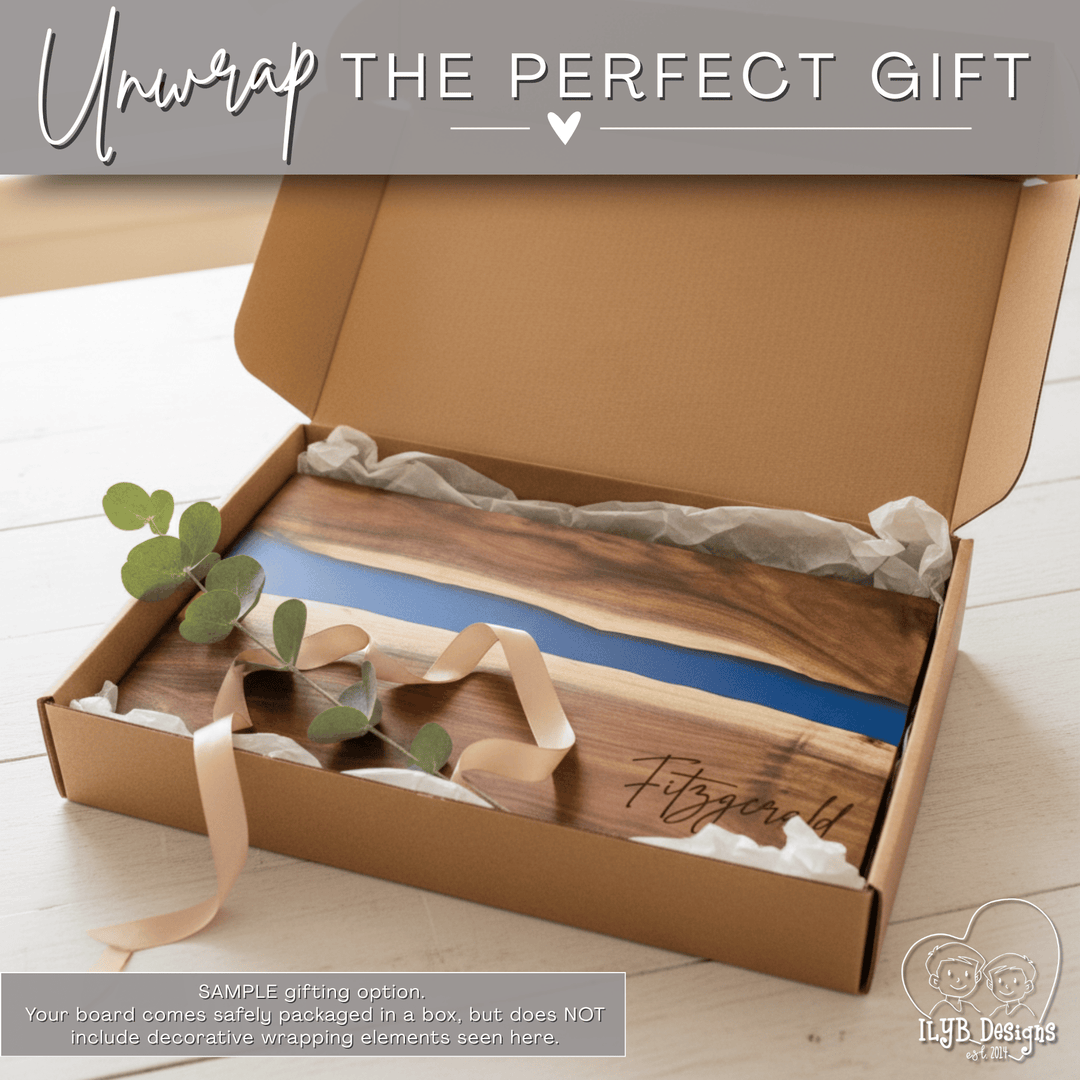 Wood charcuterie board with blue resin inlay and name engraved in bottom right corner in an open gift box. Text on top of photo says 'unwrap the perfect gift'. Bottom text says 'Sample gift option. Your board comes safely packaged in a box, but does not include decorative wrapping elements seen here.'
