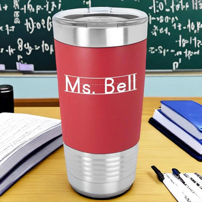 Red 20 ounce Polar Camel stainless steel tumbler with silicone sleeve that is laser engraved with Ms. Bell in a white font the looks like primary lined writing paper. Teacher tumbler is sitting on a desk with school supplies.