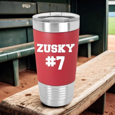 Red 20 oz Polar Camel stainless steel tumbler with a red silicone sleeve laser engraved in white with a personalized name and sports team number in a sporty font. Tumbler is sitting on bench with baseball dugout behind it.