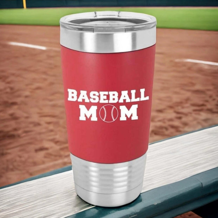 Red 20 oz Polar Camel stainless steel tumbler with a red silicone sleeve laser engraved in white that says baseball mom. The O of mom is a baseball. Tumbler is sitting on bleacher with baseball field behind it.