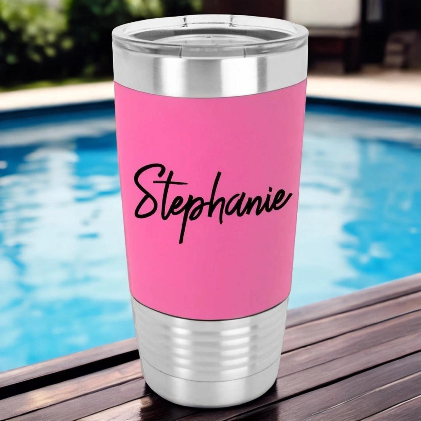 Stainless steel tumbler with pink silicone sleeve laser engraved in black with a personalized name in a modern script font. Drinking tumbler is sitting on a table in front of a pool.