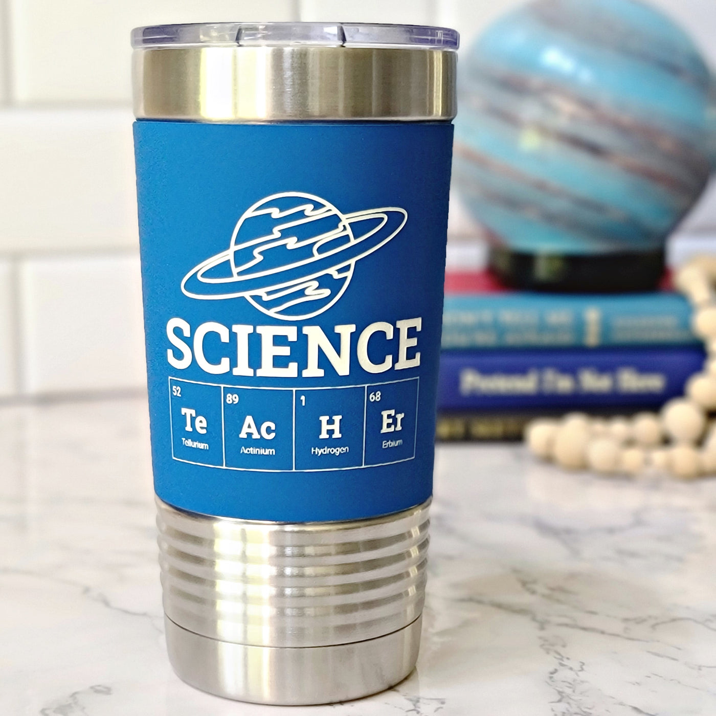 Blue stainless steel tumbler with white design laser engraved that has a planet on top of the word science and the periodic table spelling teacher underneath.