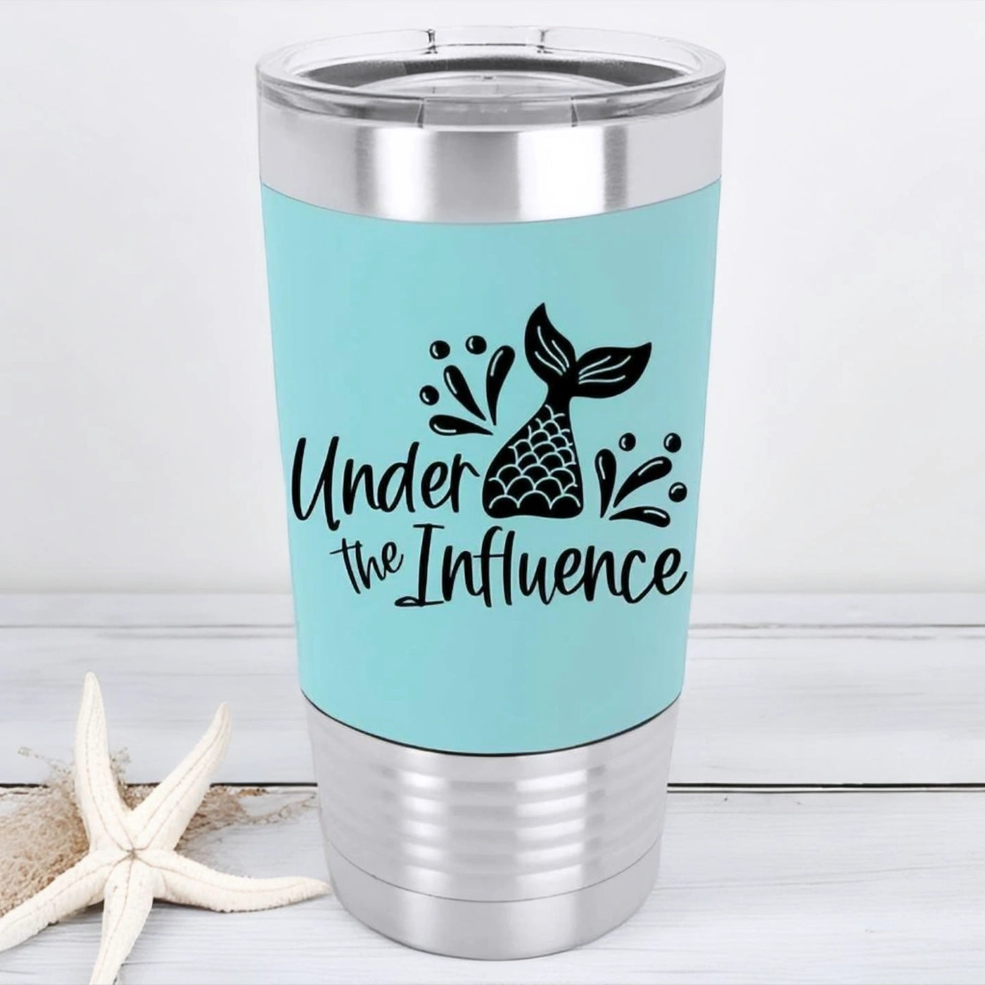 Teal 20 oz stainless steel tumbler with silicone sleeve laser engraved in black and says under the influence with a mermaid tail and water splash drops.