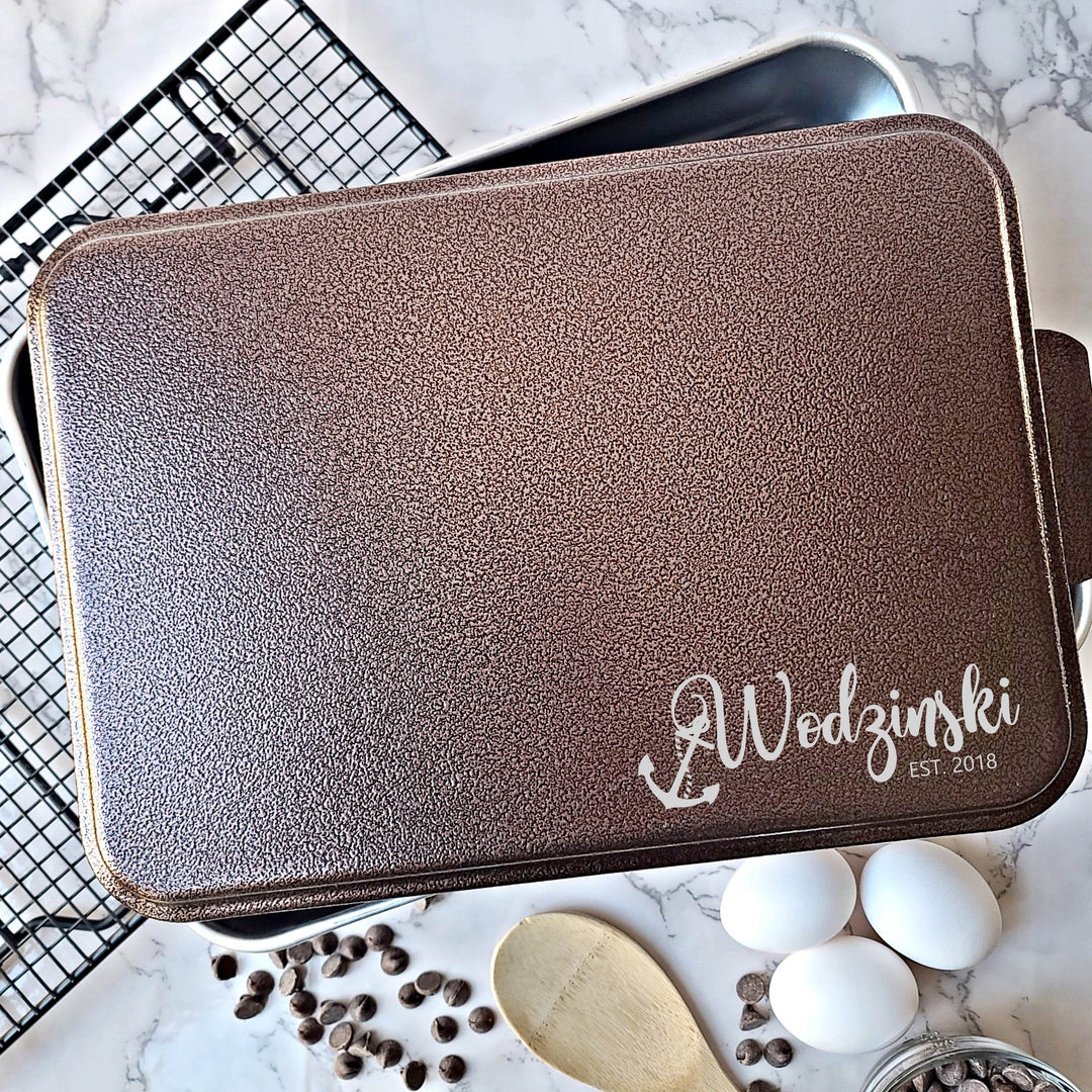 Metal casserole dish with copper lid engraved with a personalized name and established date with anchor hanging from first letter of name on a kitchen counter with eggs and spices.