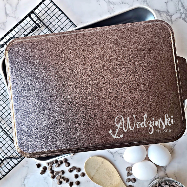Metal casserole dish with copper lid engraved with a personalized name and established date with anchor hanging from first letter of name on a kitchen counter with eggs and spices.