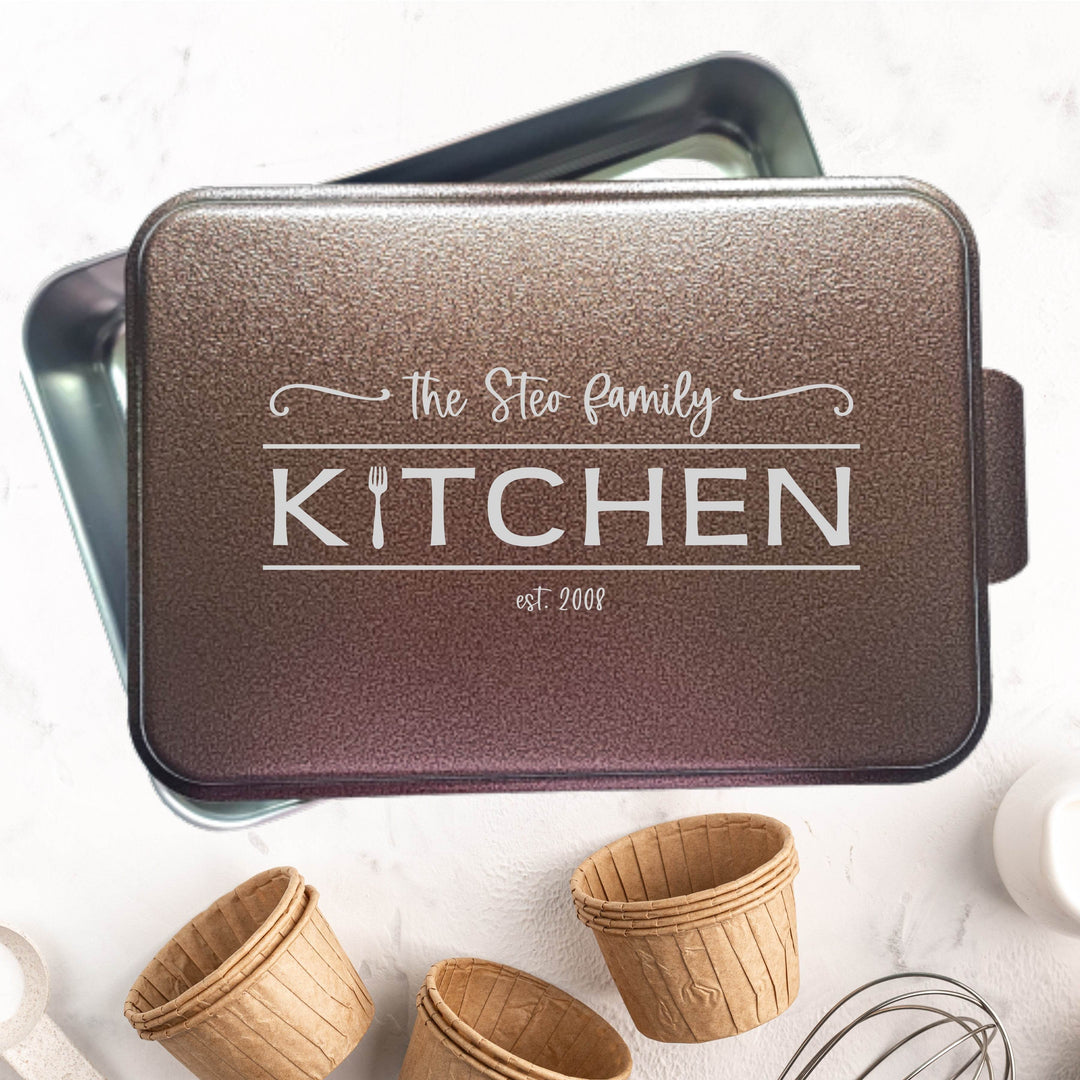 Metal baking pan with 'The Steo family KITCHEN est. 2008' text engraved on a copper powder coated lid, surrounded by small cupcake liners on a light background.
