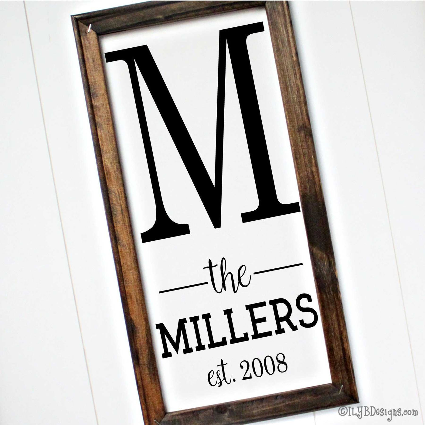 A 10"x20" dark walnut stained frame on a white canvas with a black design placed vertically. The design is a large printed initial letter on top with the word "the" placed in between 2 lines followed by a last name in a print font followed by an established year underneath.